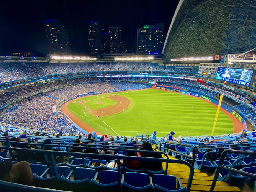 Aerial view of a night baseball game in a packed stadium with city skyline.